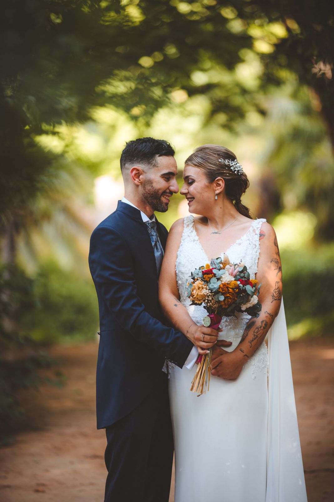 Los novios se miran sonriendo bajo los árboles, ella con un ramo de flores secas