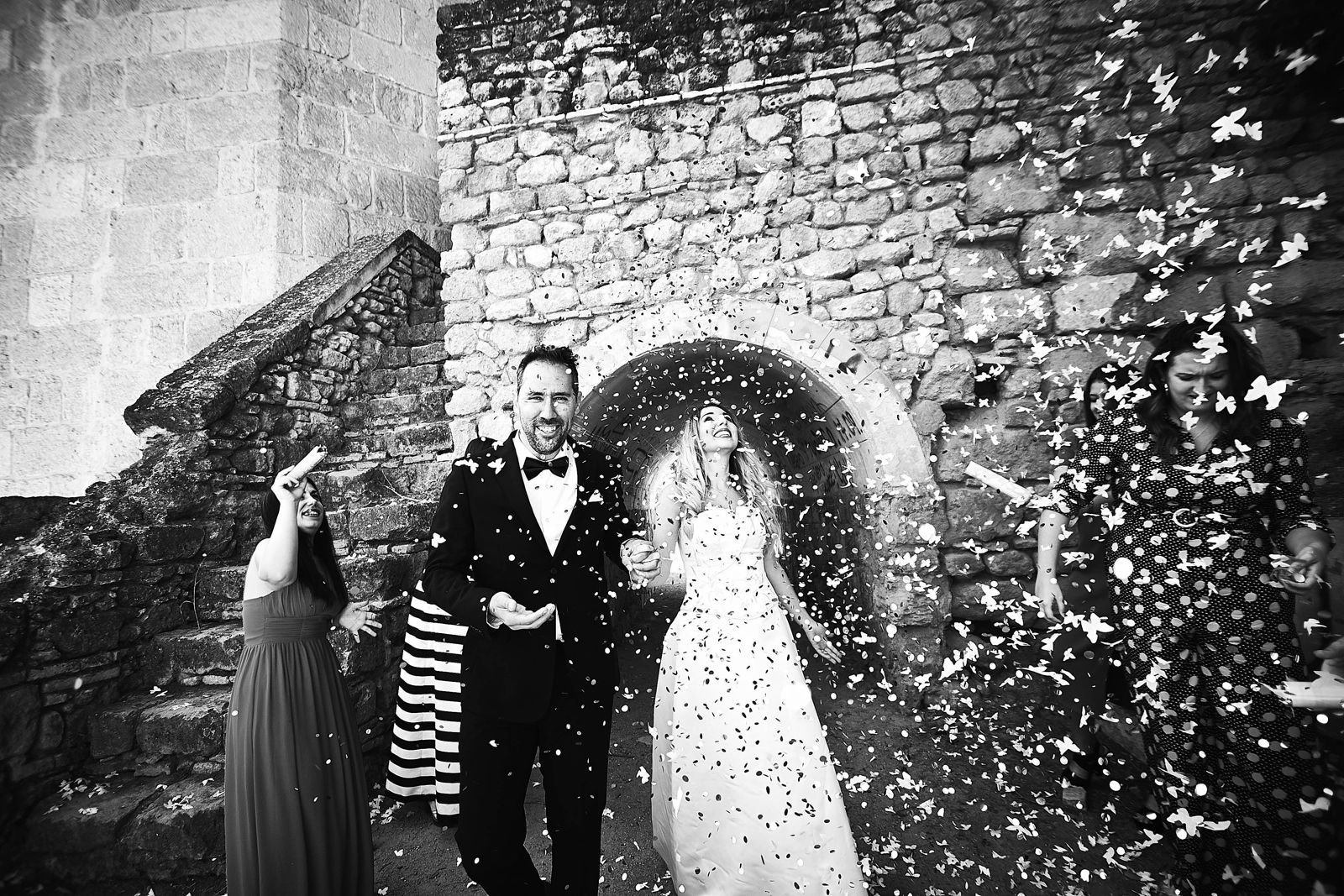 Los novios salen de la ceremonia bajo una lluvia de confeti junto a un muro de piedra